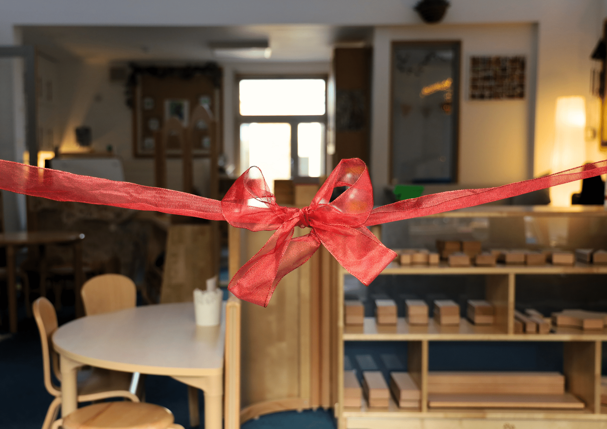 A large red ribbon tied in a bow stretches across the entrance to a well-lit classroom with wooden furniture and shelves filled with wooden blocks.