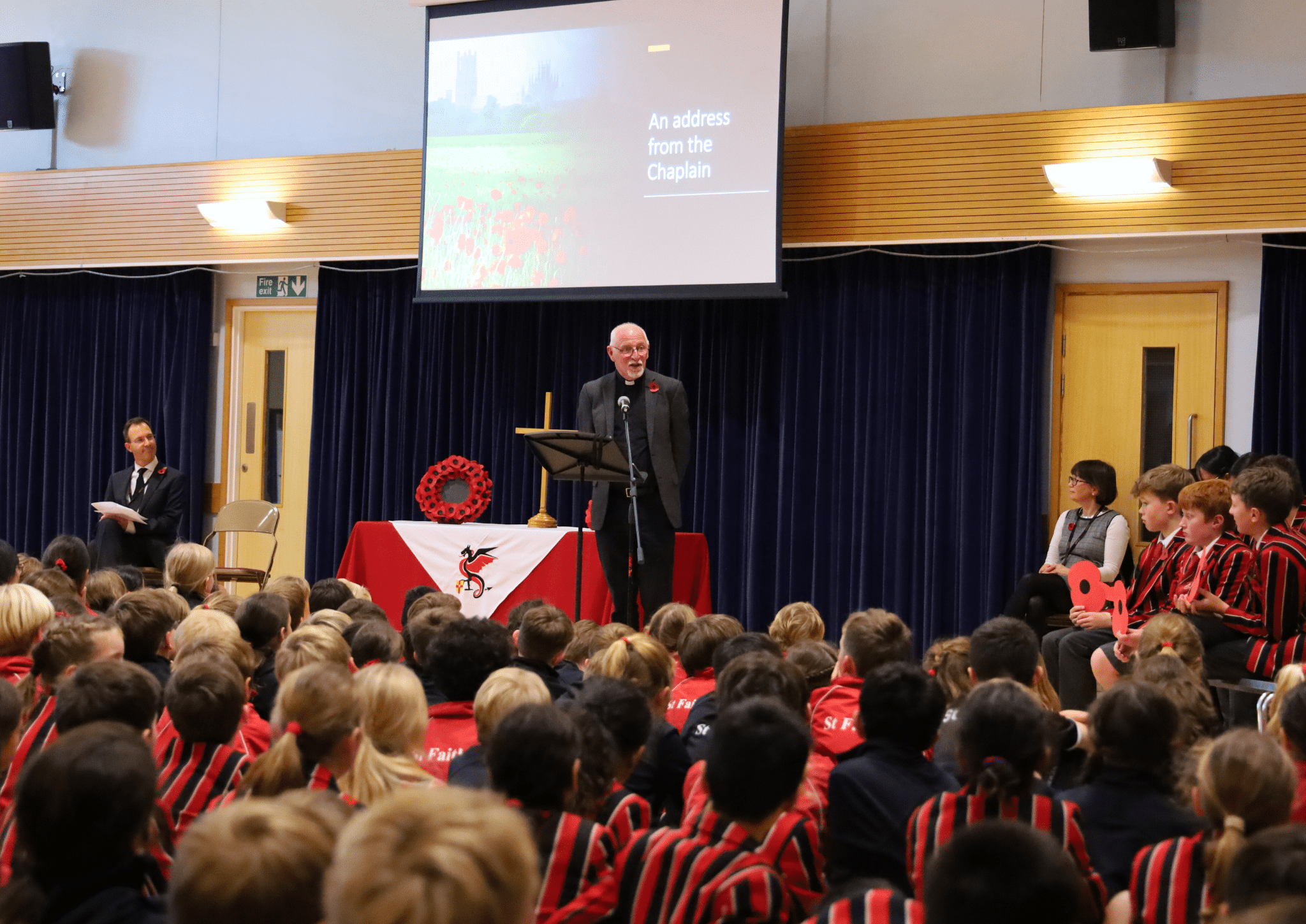 A chaplain speaks at a podium in a school assembly hall, addressing students in red and black striped uniforms. A screen behind him reads An address from the Chaplain. Faculty sit nearby, and students listen attentively.