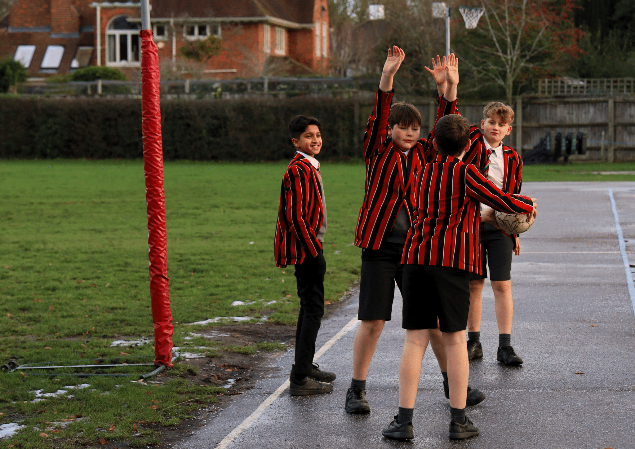 Four boys in red and black striped blazers stand on a wet outdoor basketball court. Two boys face each other, one holding a ball, while another raises his arms. A fourth boy smiles, looking toward the camera.