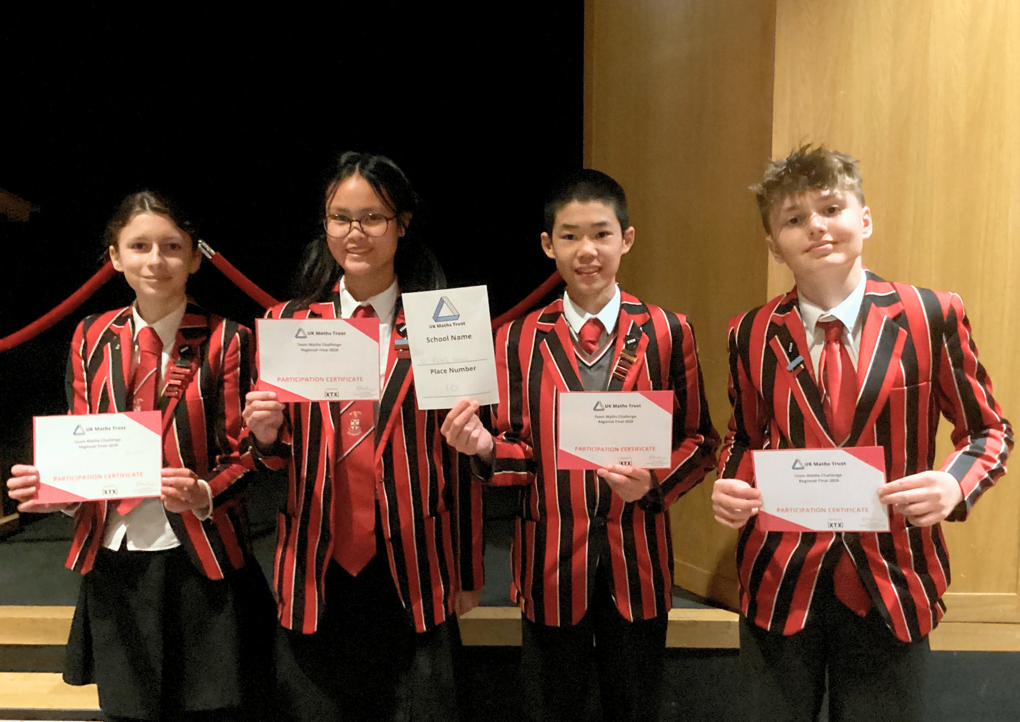Four students in red and black striped blazers stand indoors, smiling and holding participation certificates and a sign reading “School Award.” They are positioned side by side in front of a wooden backdrop.