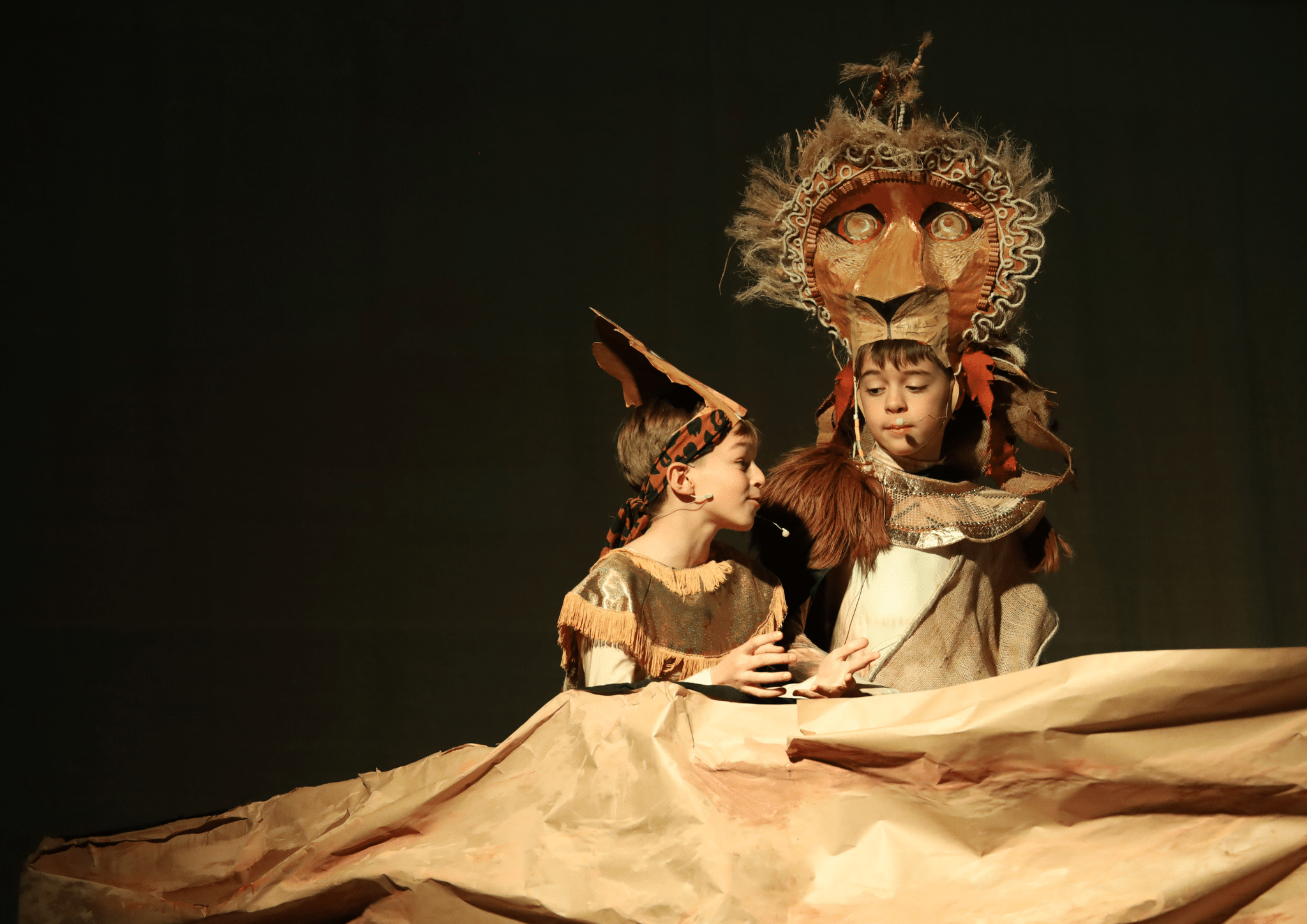 Two children in animal costumes, one as a lion and the other as another animal, perform on stage with a large sheet of brown paper representing the landscape. The scene is dramatically lit against a dark background.