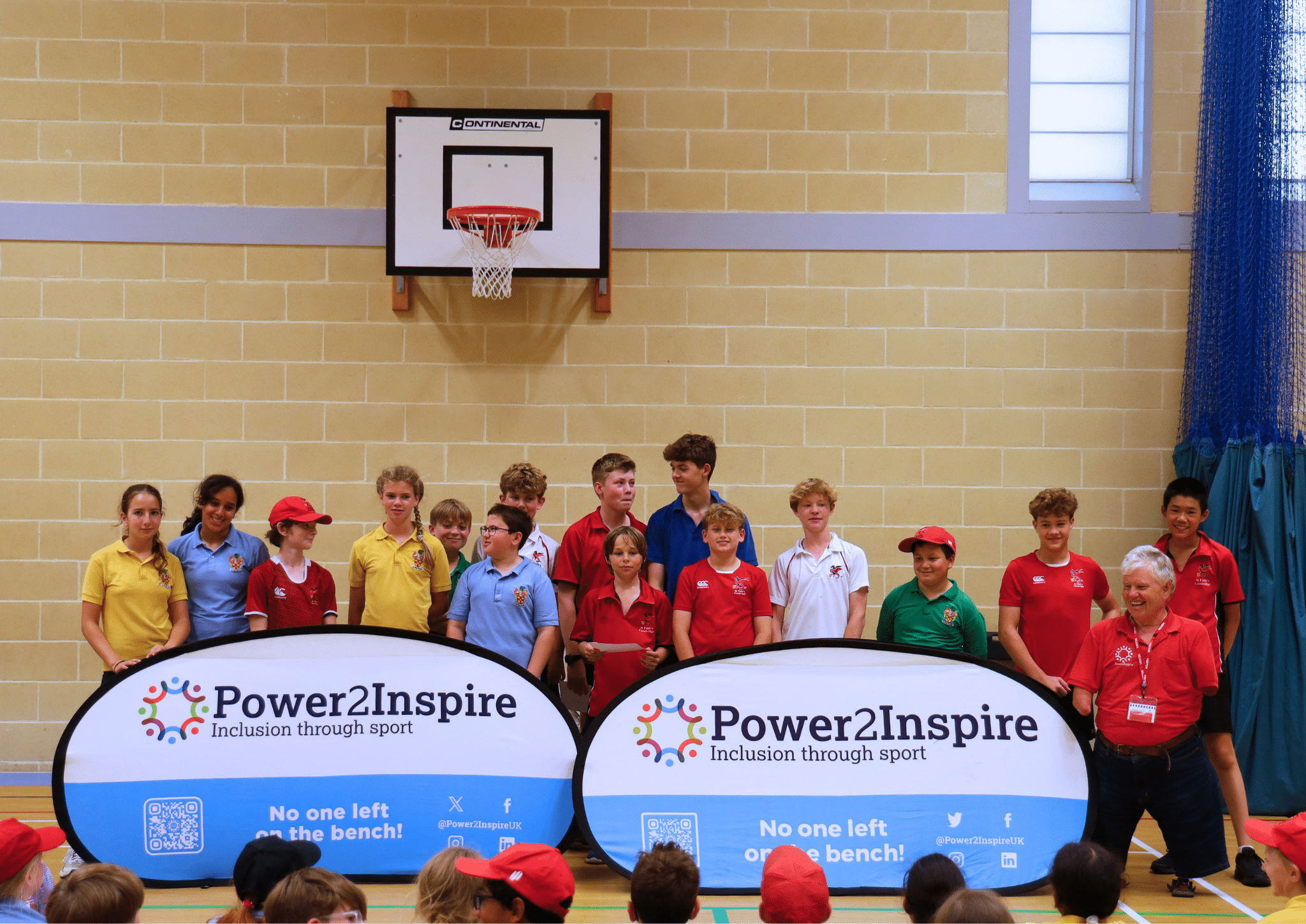 A diverse group of children and two adults stand in a sports hall, smiling behind two banners reading “Power2Inspire: Inclusion through sport.” Some children wear coloured shirts and hats, and a basketball hoop is visible behind them.
