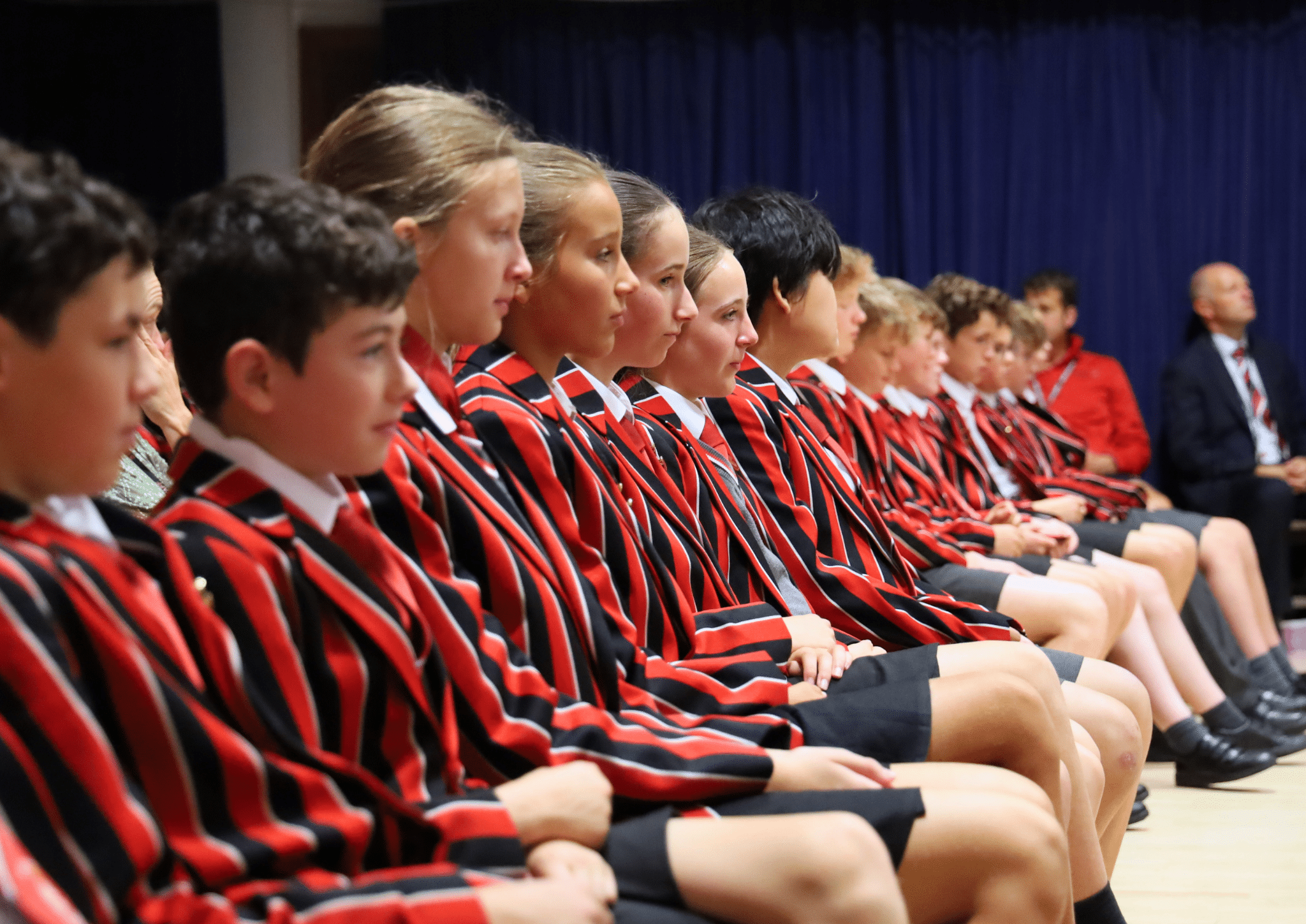 A row of students in red and black striped blazers and shorts sit attentively in a hall, with a dark blue curtain in the background. An adult sits at the end of the row.