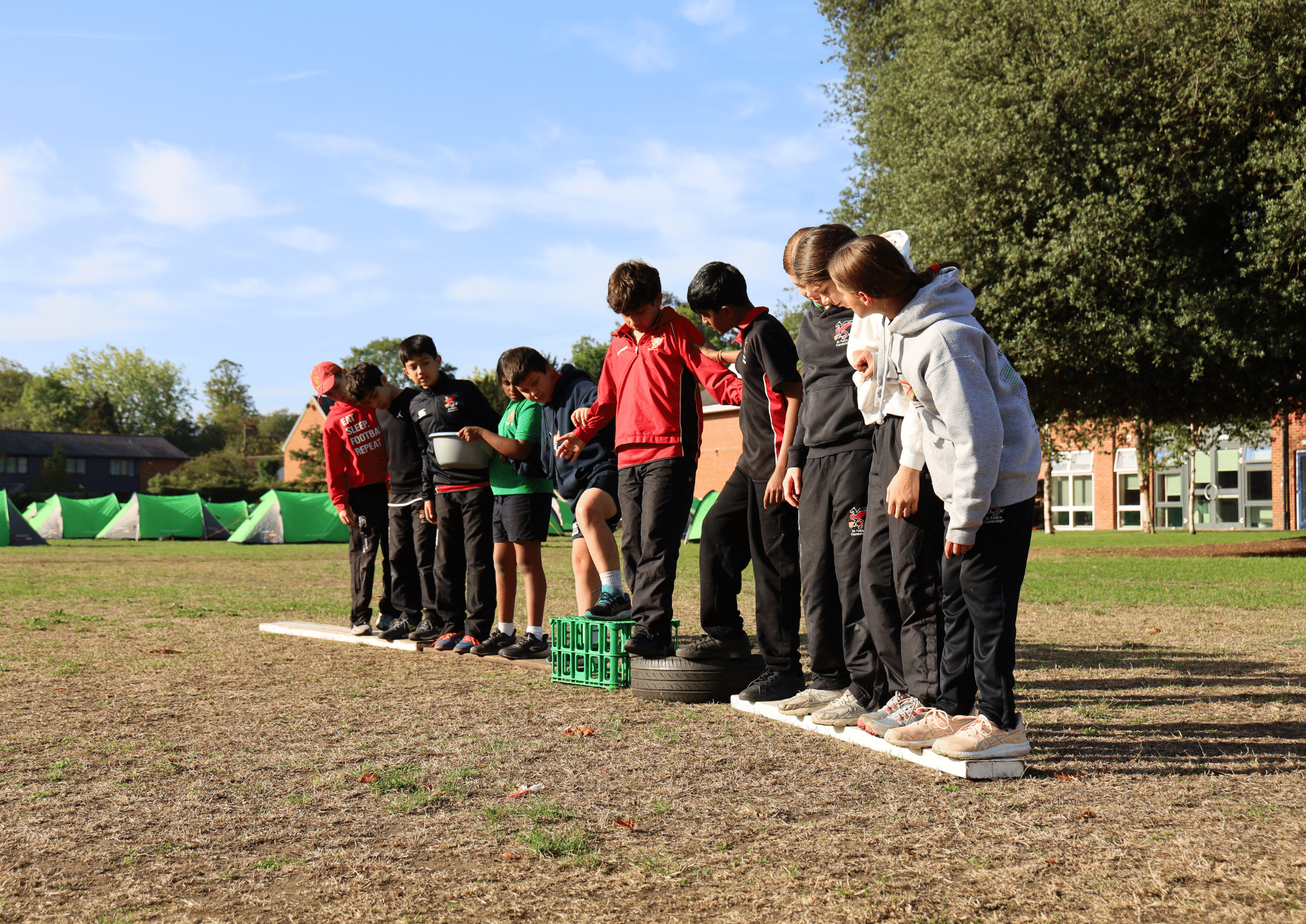 A group of children in sports kit balance on crates, tyres, and boards outdoors, working together on a grassy field under a sunny sky, with trees and buildings in the background.