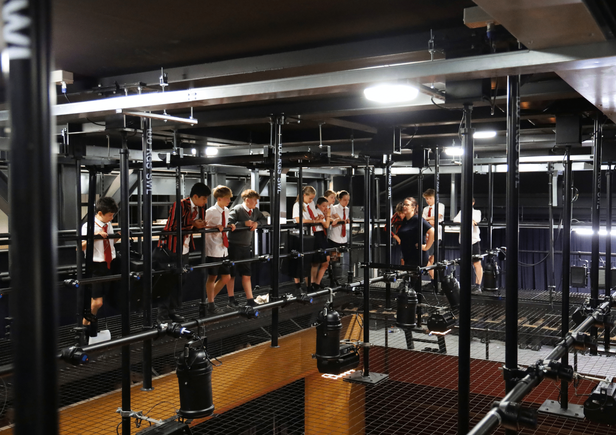 A group of students in uniforms walks on a metal walkway above a theatre stage, surrounded by lighting equipment and beams.
