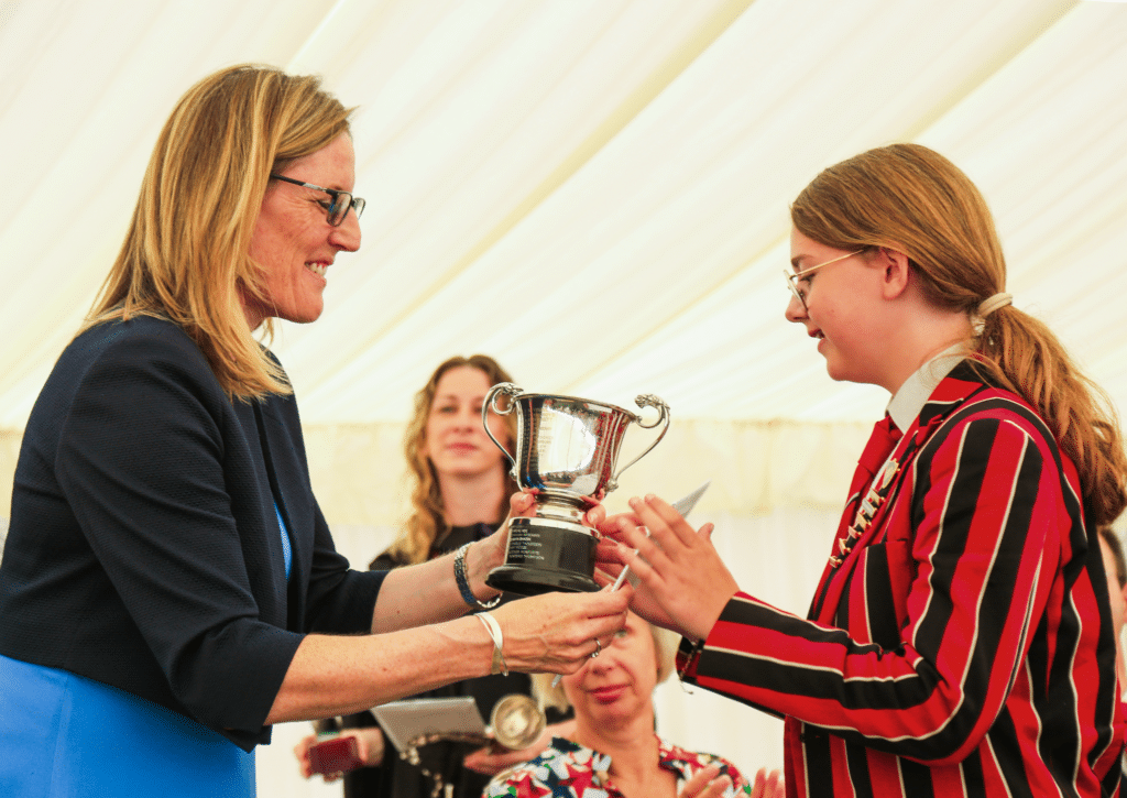 A woman hands a silver trophy to a young person in a red and black striped blazer, whilst others watch under a white marquee.