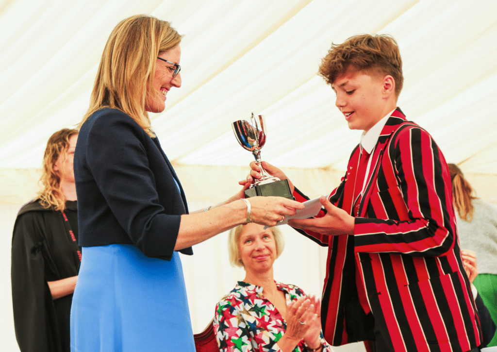 A woman hands a silver trophy to a smiling young person in a red and black striped blazer, whilst people in the background watch and applaud under a white marquee.
