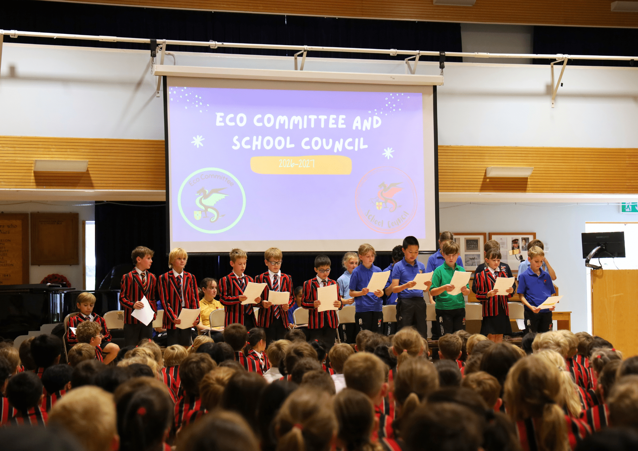 Students in school uniforms stand on a stage holding papers in front of a projection screen titled Eco Committee and School Council 2026–2027.