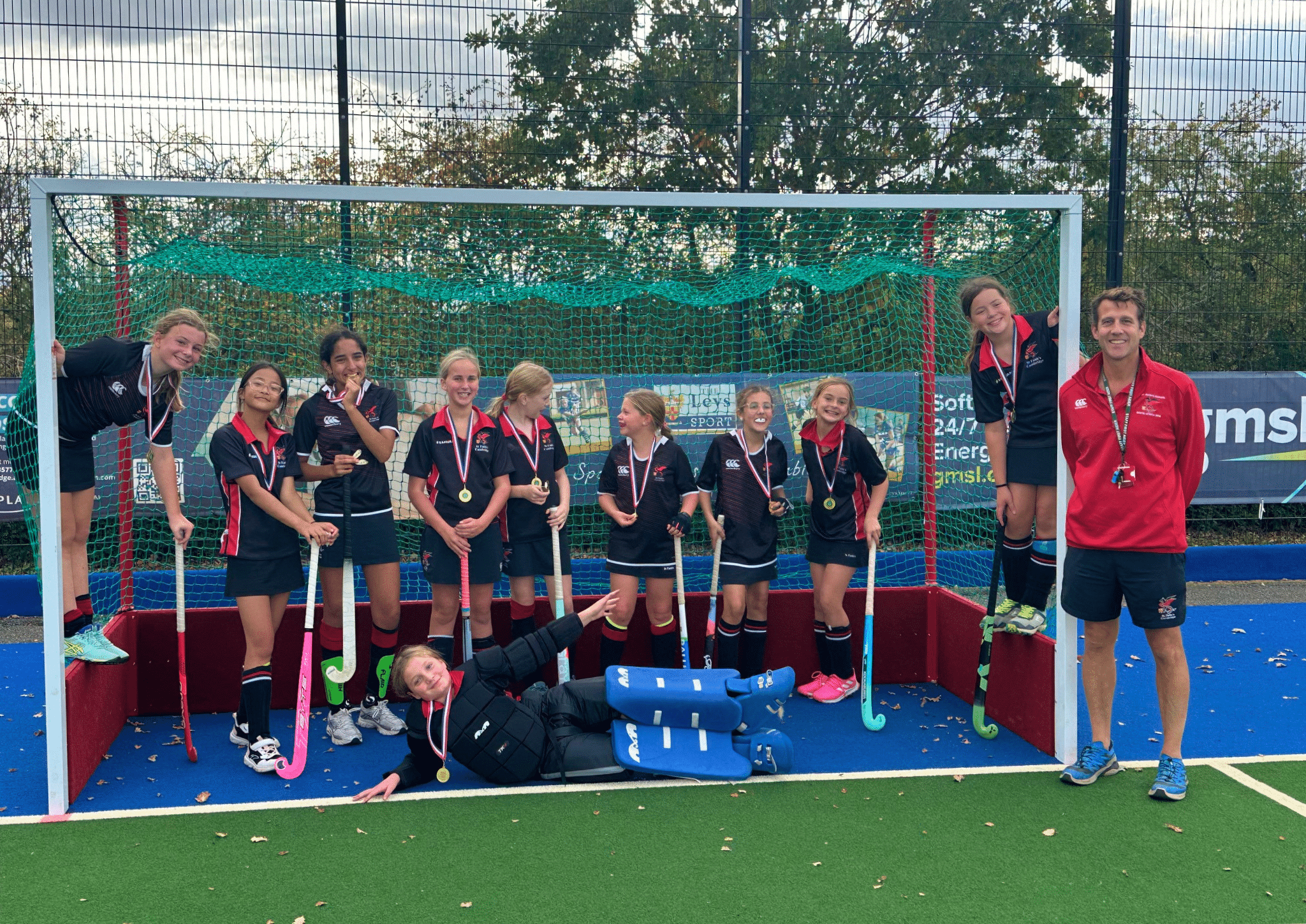 A youth hockey team and their coach pose for a photo in front of a goal on a blue pitch, all wearing medals.