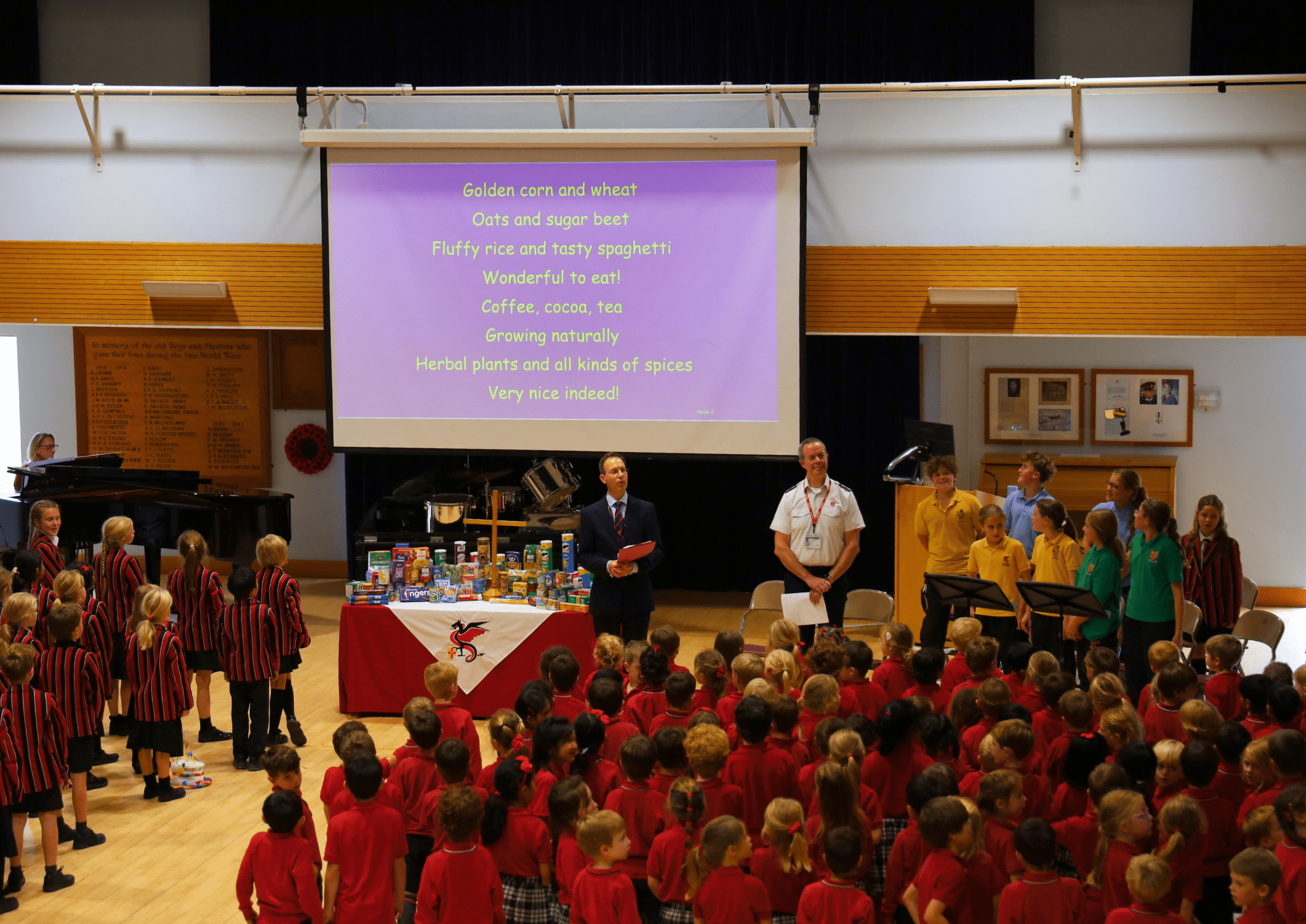 A man in a suit and a man in a uniform stand before a stage of schoolchildren, with a projector screen displaying a song about crops and food above a table of groceries.